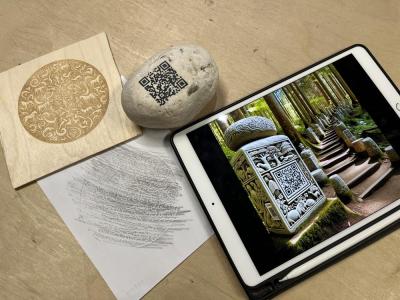 three objects are shown; a square piece of wood with a circular drawing with multiple animals, a rock with a qr code and a tablet with the image of a stone totem with a qr code